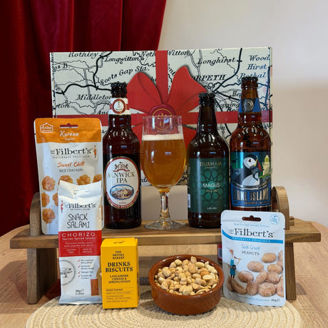 A curated gift basket or snack and beer selection arranged on a small wooden stand in front of a map-style gift box with a red ribbon. Visible items include three dark beer bottles, a filled beer glass, a bag of Kretae Sweet Butter Rice Cracker, a bag of Kretae Chorio Salt, a box of Drinks Snacker, and a bag of Tilbert's Salt Crust Peanuts, with a bowl of biscuits in front.
