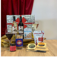 A gourmet gift hamper arranged on a wooden table with a red curtain backdrop. The set includes bottles of wine, two filled wine glasses, a jar of preserves, a bag of cheddar cheese, a block of Brie cheese, a bag of Nutella, a bag of Filbert's chocolate chip cookies, and a blue box of crackers, all set against a decorative map-patterned gift bag.
