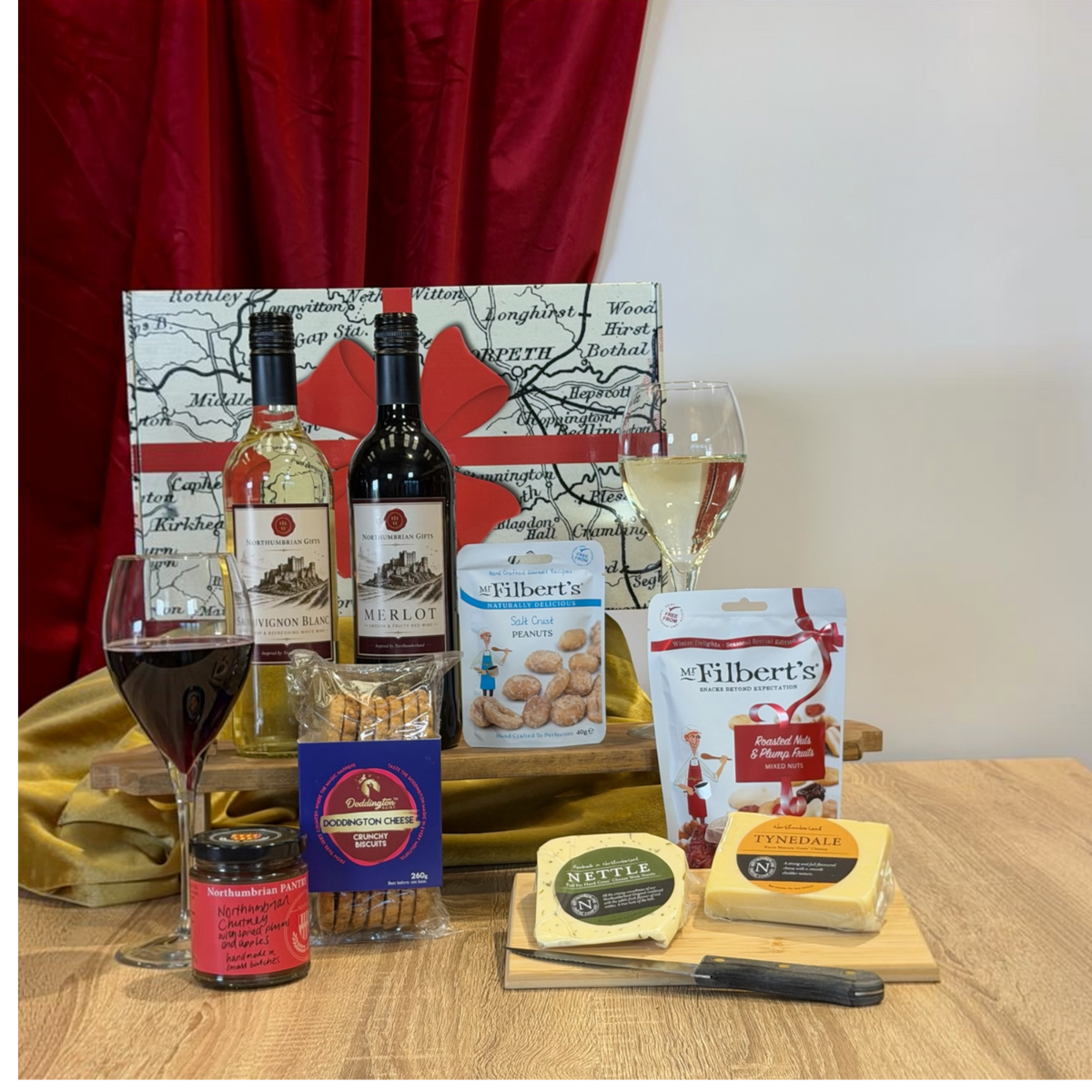 A gourmet gift hamper arranged on a wooden table with a red curtain backdrop. The set includes bottles of wine, two filled wine glasses, a jar of preserves, a bag of cheddar cheese, a block of Brie cheese, a bag of Nutella, a bag of Filbert's chocolate chip cookies, and a blue box of crackers, all set against a decorative map-patterned gift bag.