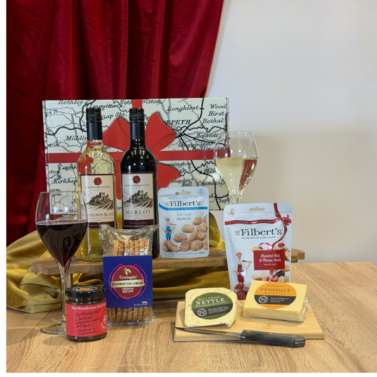 A gourmet gift hamper arranged on a wooden table with a red curtain backdrop. The set includes bottles of wine, two filled wine glasses, a jar of preserves, a bag of cheddar cheese, a block of Brie cheese, a bag of Nutella, a bag of Filbert's chocolate chip cookies, and a blue box of crackers, all set against a decorative map-patterned gift bag.