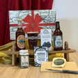 A curated gift set arranged on a wooden stand and tabletop, featuring two brown beer bottles, a pint glass, a small jar, a cookbook, a map-style sign, and a cheese wheel with a knife, set against a red curtain and cream wall.