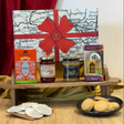 A curated gift hamper arrangement featuring a large map-patterned gift box with a red ribbon and bow, alongside boxes and jars of food items including tea, jam, and scones, displayed on a wooden stand with decorative fabric and a plate of cookies in front.