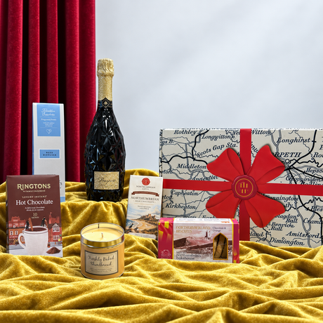 “Northumbrian pamper hamper with Prosecco, chocolate, hot chocolate, biscuits, candle and reed diffuser beside a gift box.”